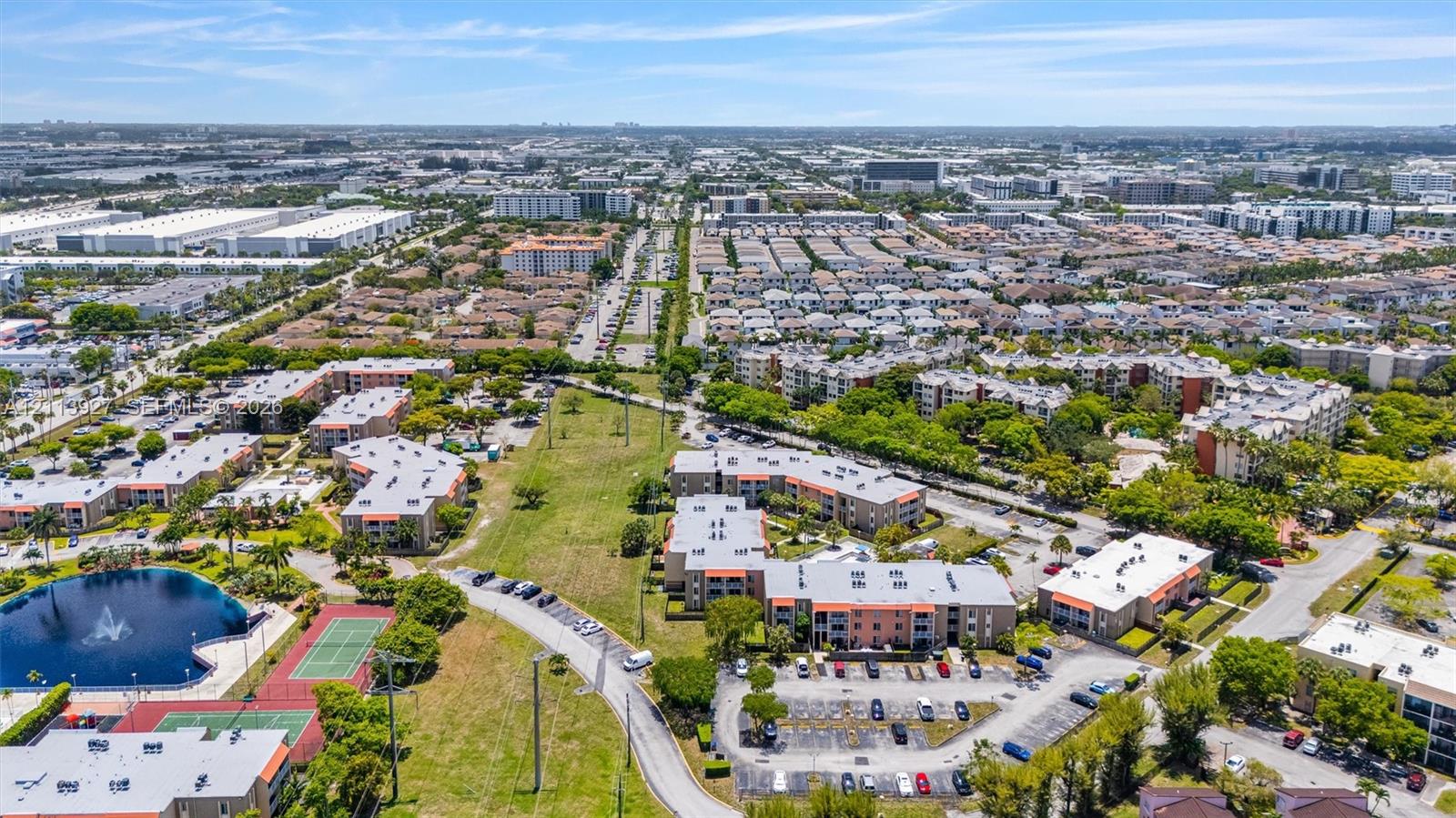Aerial perspective featuring multi-story residential buildings, a central water feature with a fountain, paved surface parking, and tennis courts