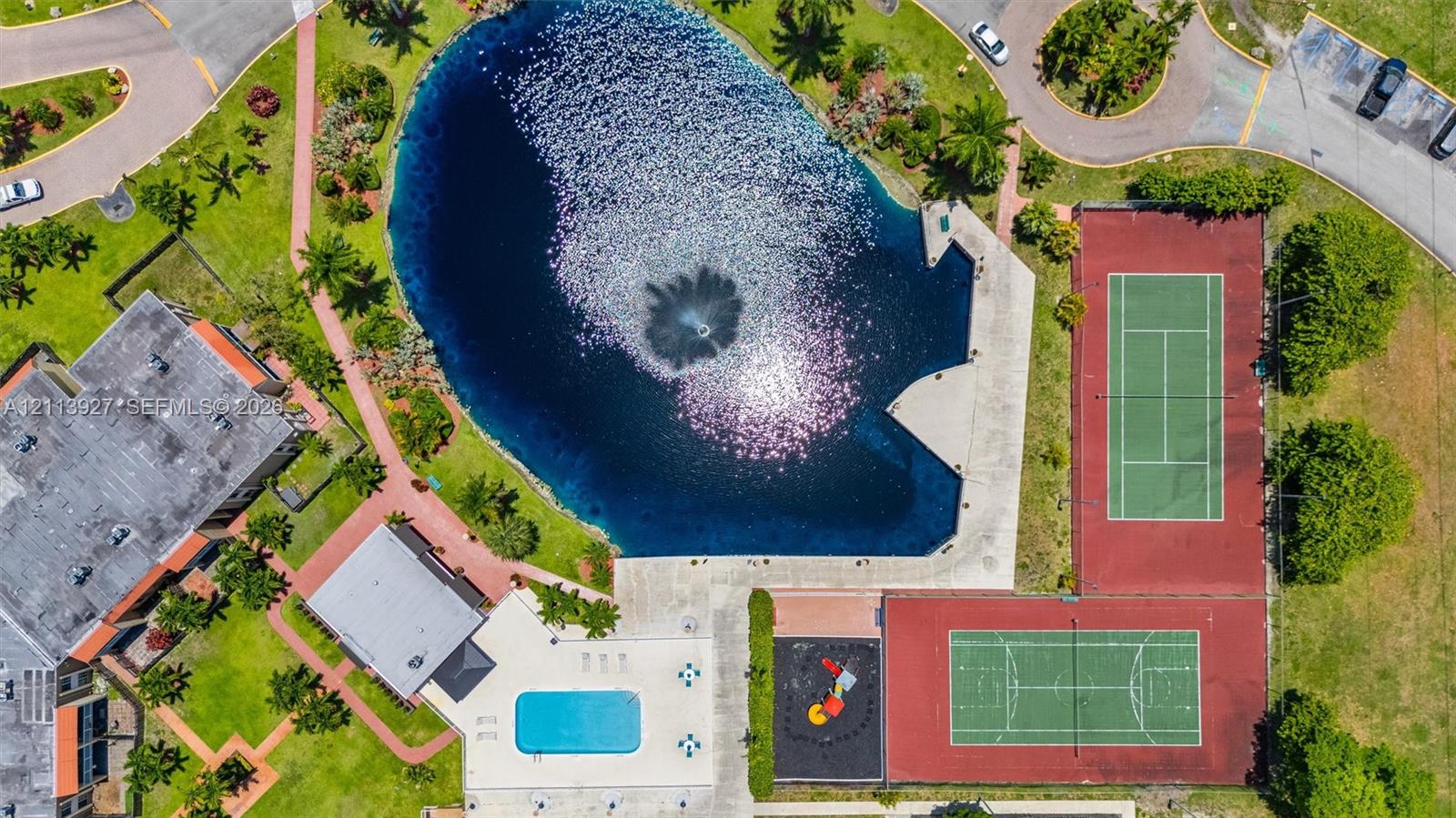 Aerial view featuring a central lake with a fountain, swimming pool, two hard-court surfaces, and paved walkways throughout the grounds