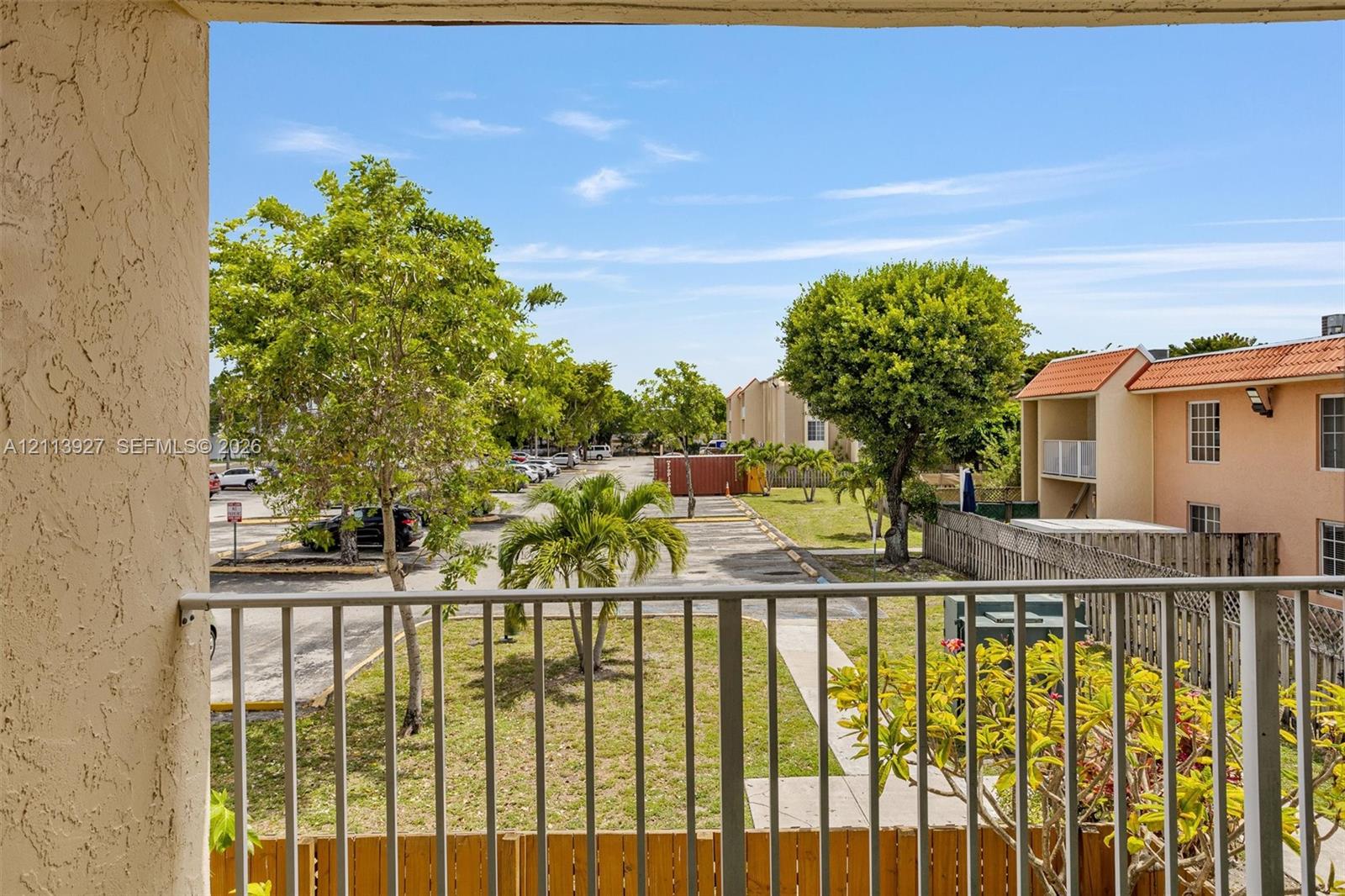 Balcony featuring metal railing and stucco pillar finish, overlooking exterior grounds with mature landscaping and paved parking area