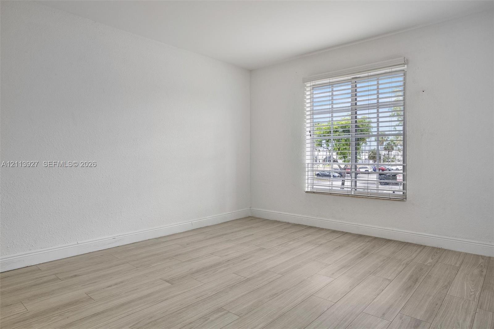 Neutral interior space featuring wood-finish flooring, crisp white walls, and a large window equipped with horizontal slat blinds