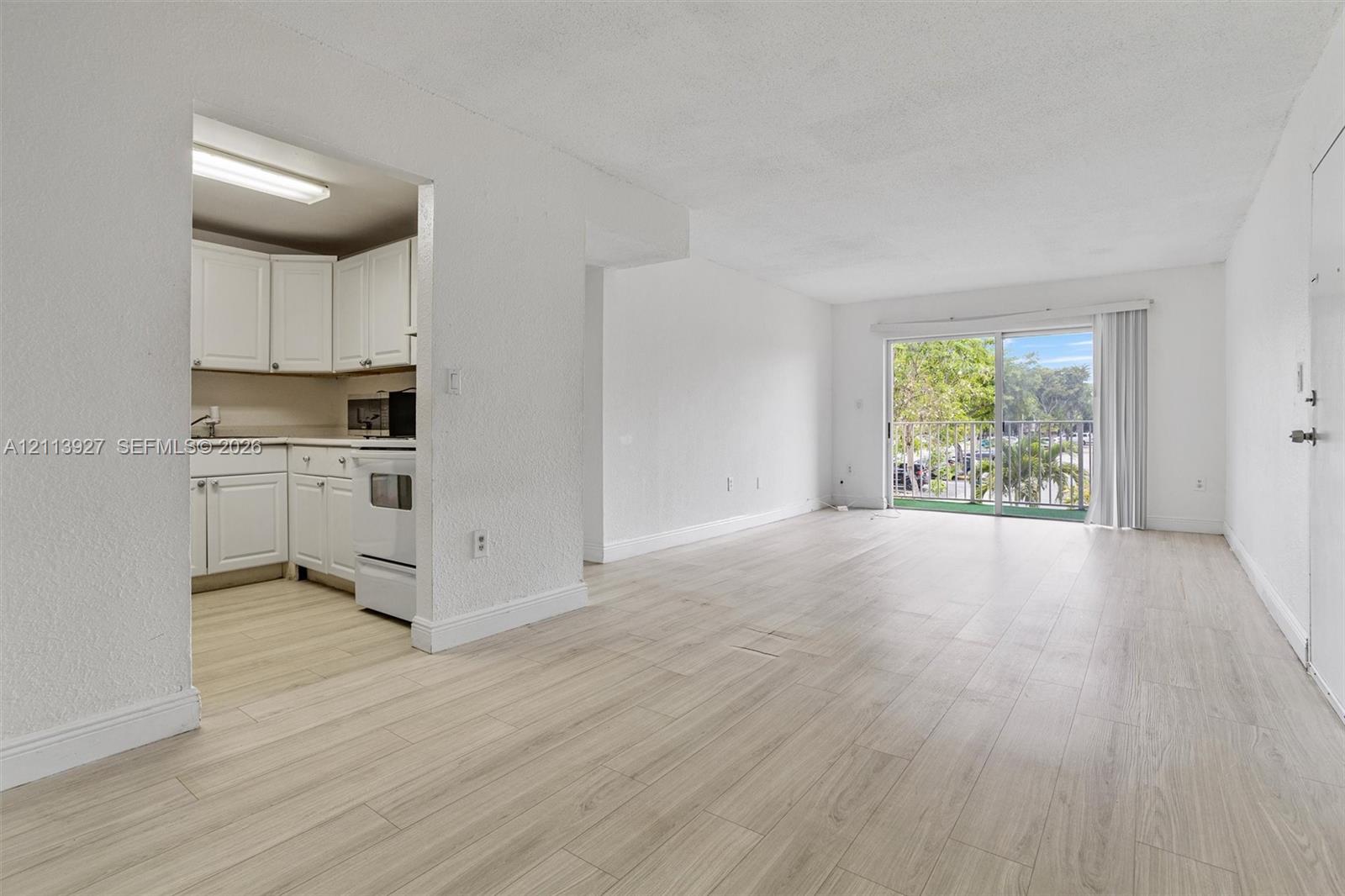 Open-concept interior featuring wood-finish flooring, sliding glass door with access to an exterior balcony, and kitchen with white cabinetry and flush-mount lighting