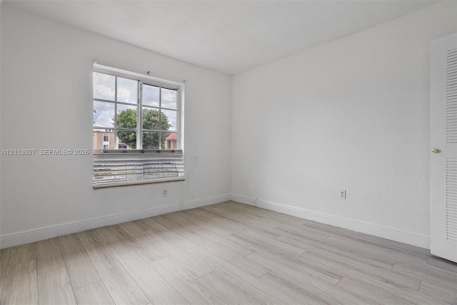 Neutral interior featuring light wood-finish flooring, white walls, and a multi-pane window with louvered blinds