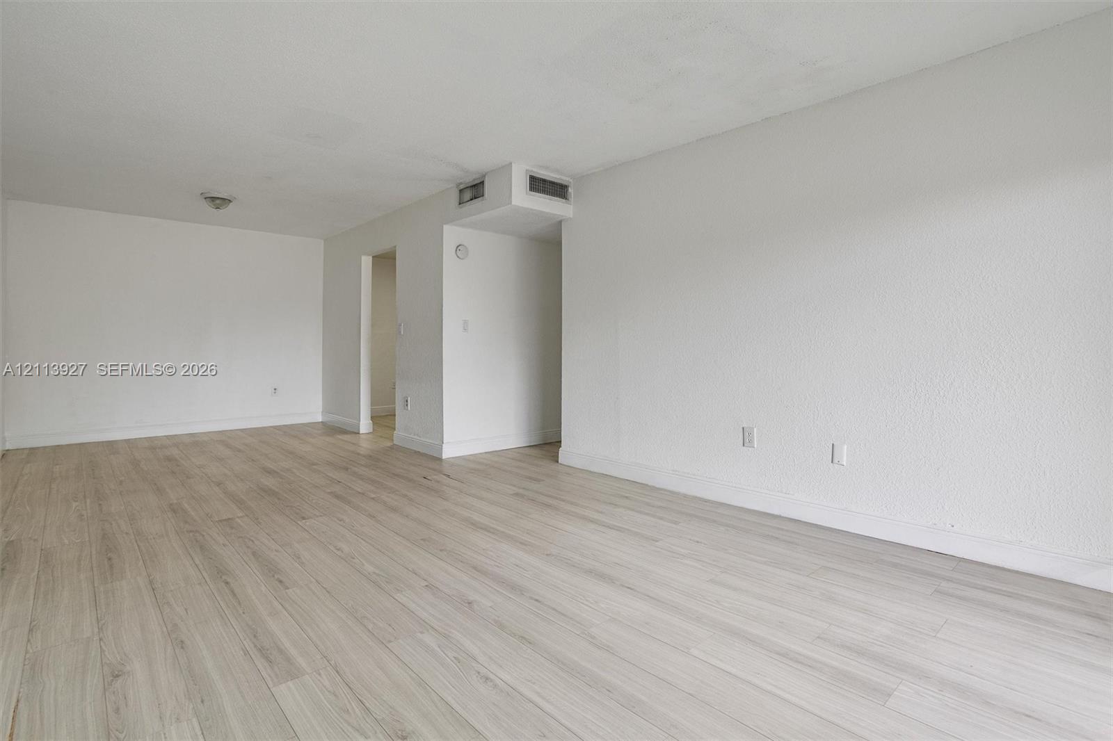 Open-concept interior featuring light-toned wood-finish flooring, smooth-textured walls, and a flush-mount ceiling light fixture