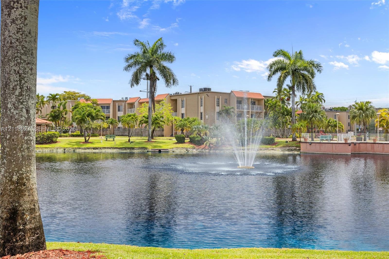 Waterfront grounds featuring a central fountain, manicured lawn, palm trees, and multi-story building exterior
