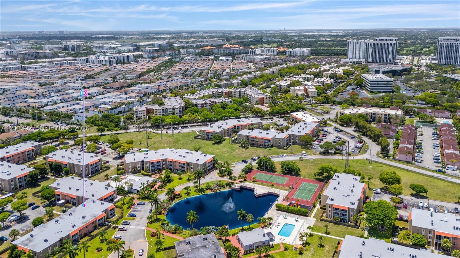 Aerial perspective of a multi-unit complex featuring a central lake with a fountain, swimming pool, and multiple tennis courts