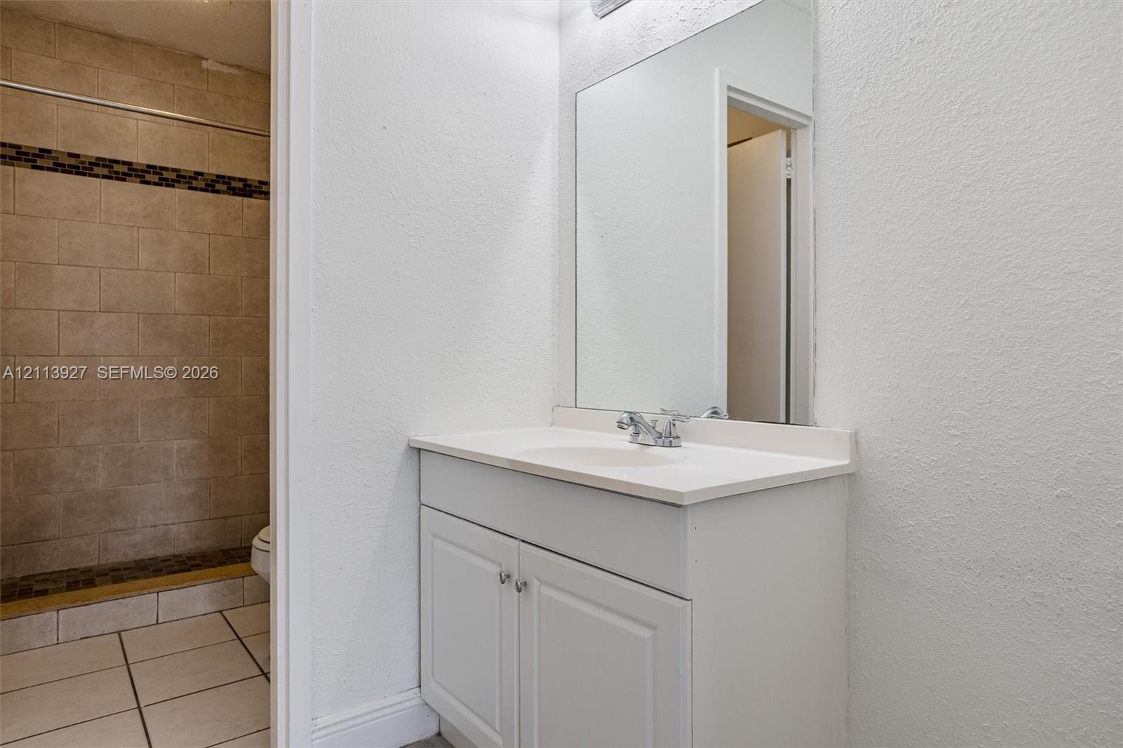 Bathroom featuring a white vanity with an integrated sink, polished chrome faucet, expansive wall-mounted mirror, and a walk-in shower with neutral stone-finish tile surround and decorative accent inlay