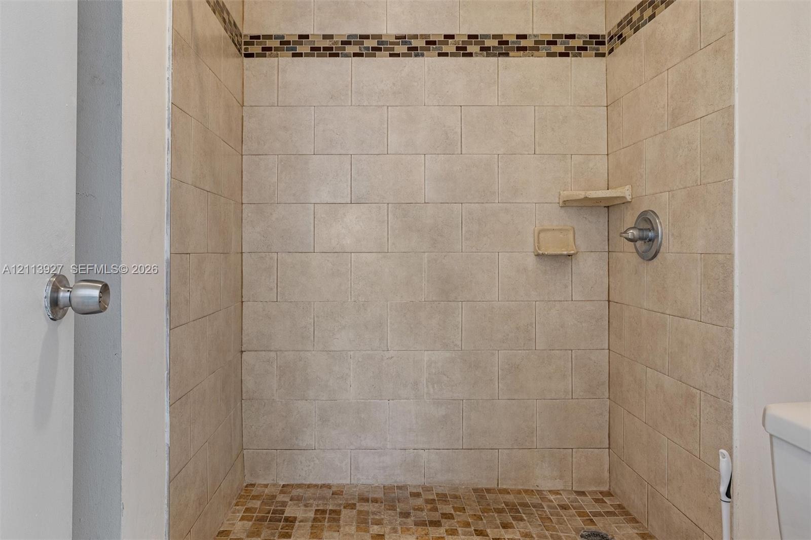 Walk-in shower featuring neutral rectangular wall tile, mosaic tile accent border, integrated corner shelving, and brushed nickel hardware