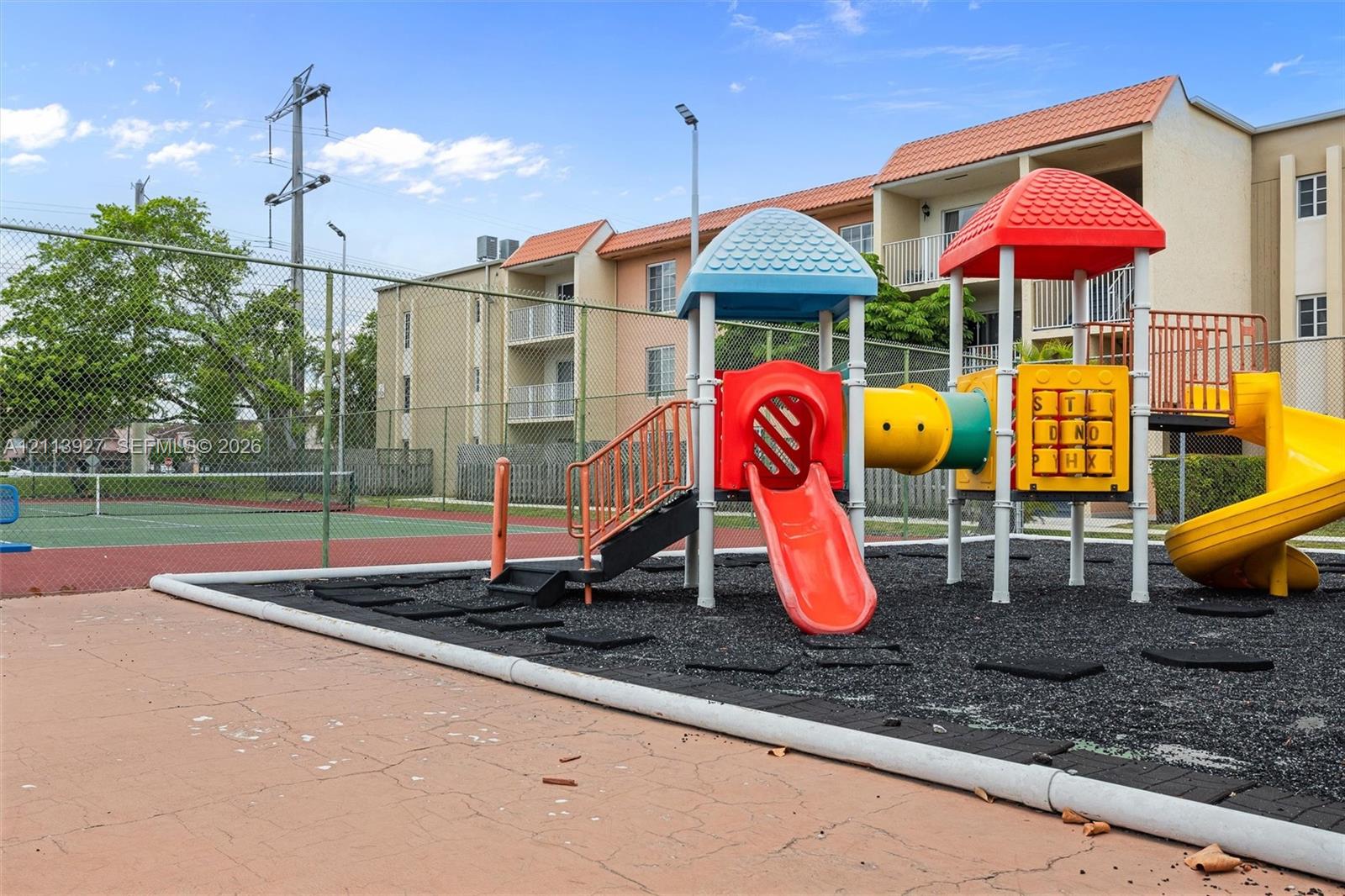 Playground area featuring a multi-tiered structure with a slide, hard-surfaced tennis court, and perimeter chain-link fencing