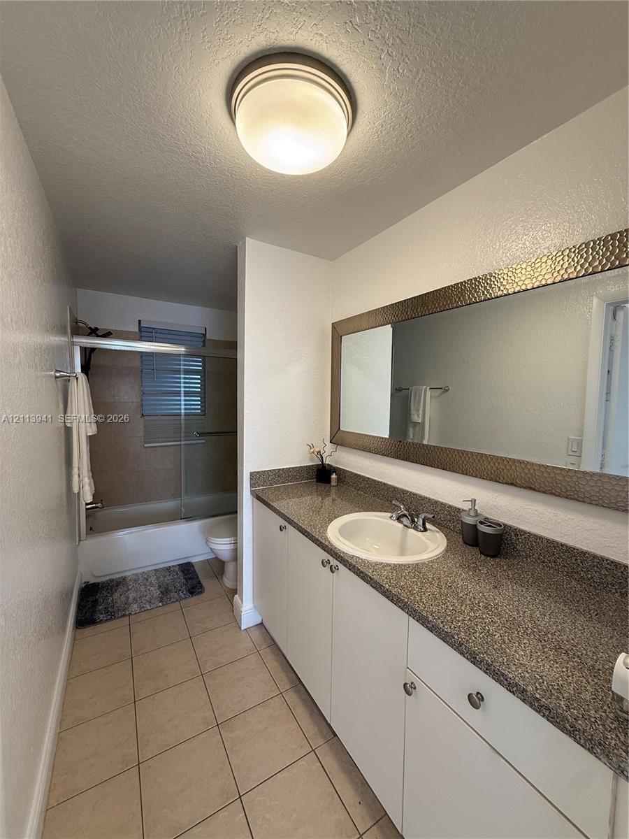 Full-width vanity with solid-surface countertop, integrated basin, tiled flooring, large framed wall mirror, and glass-enclosed shower and tub combination
