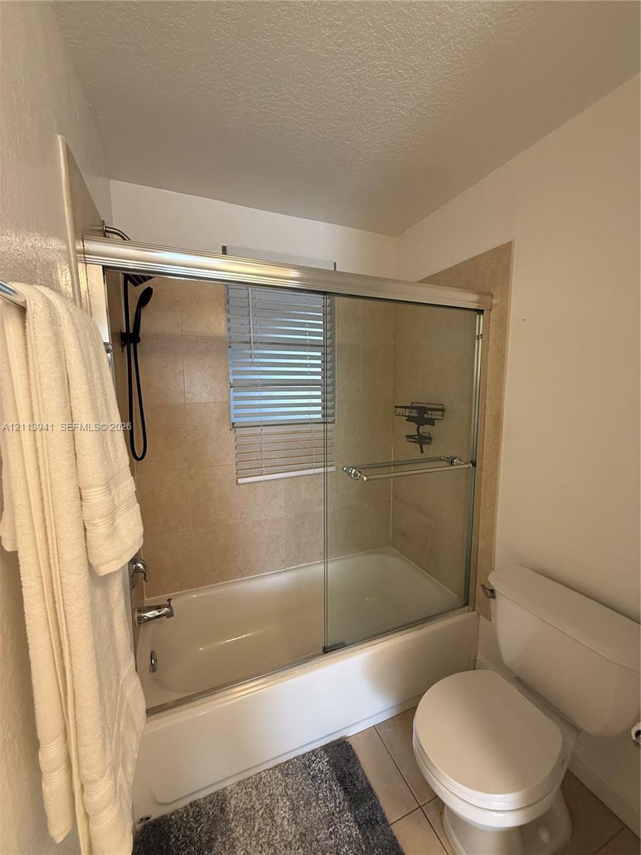 Bathtub with glass sliding doors, neutral tile surround, black handheld shower fixture, and tiled flooring