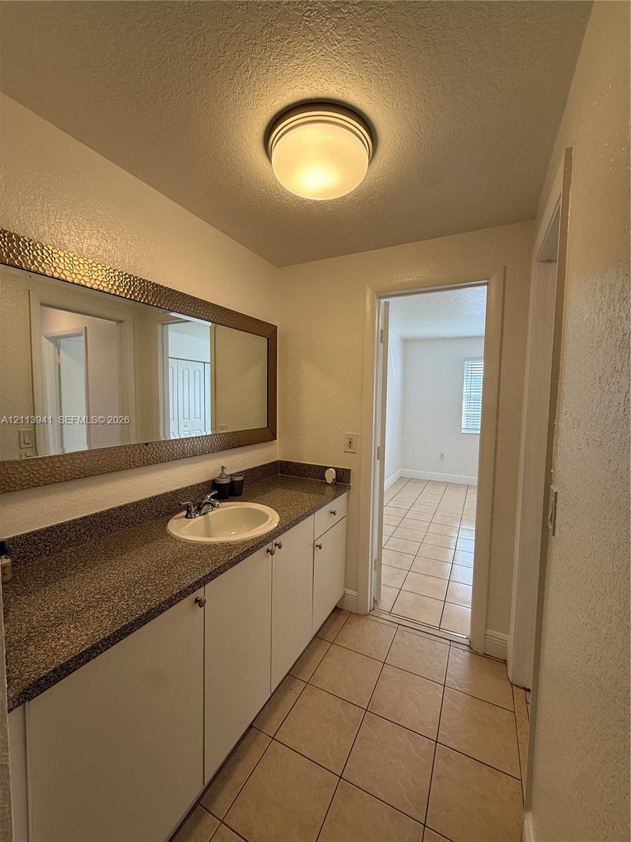 Extended vanity featuring a speckled stone-finish countertop, white cabinetry, a circular undermount sink, and a full-width mirror with a hammered metal-finish frame, complemented by ceramic tile flooring