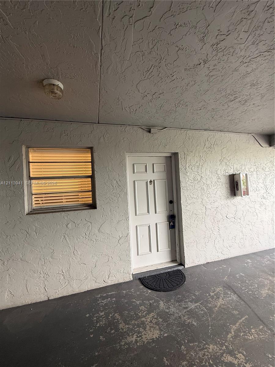 Covered exterior entryway featuring textured stucco walls, a six-panel entry door, jalousie window, and flush-mount ceiling lighting