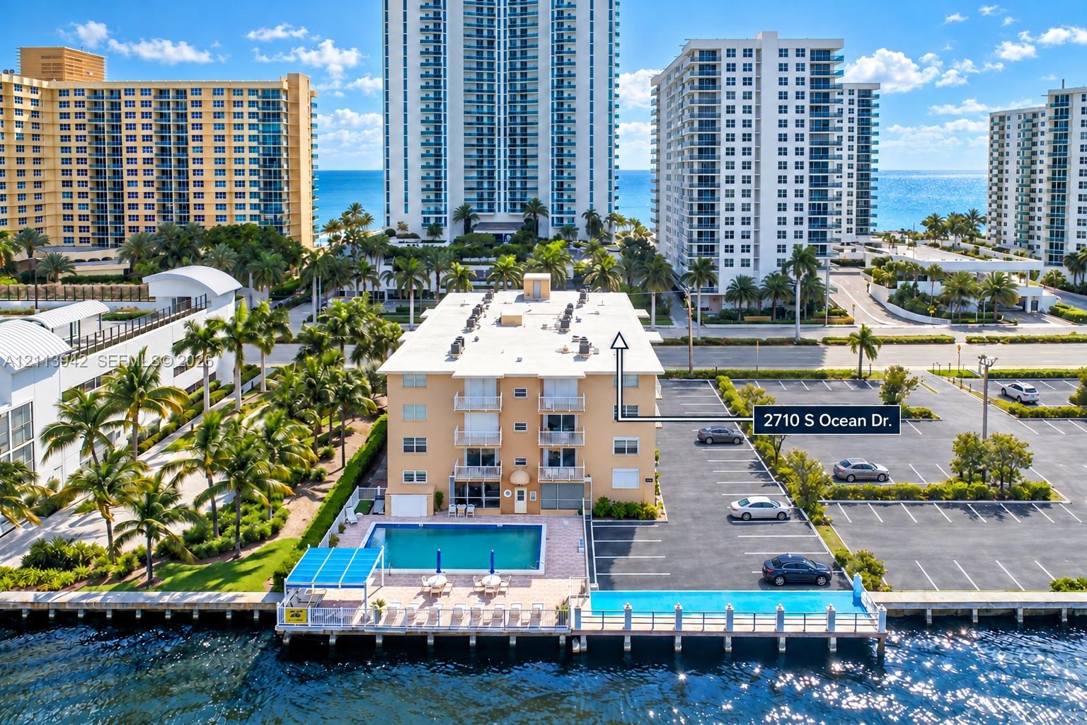 Waterfront mid-rise building featuring a ground-level swimming pool, expansive paved parking area, private dockage, and manicured palm tree landscaping