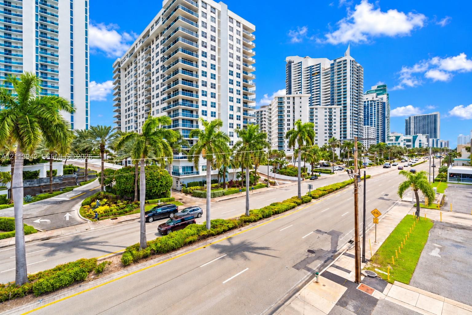 High-rise residential exterior featuring glass balcony railings, white concrete facade, mature palm tree landscaping, and paved multi-lane street access