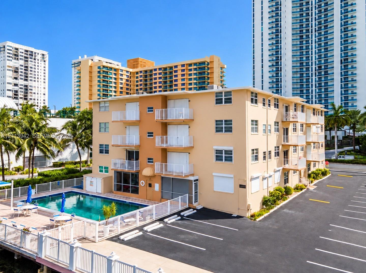 Multi-story residential building featuring private exterior balconies, an outdoor swimming pool, a perimeter fence, and surface parking