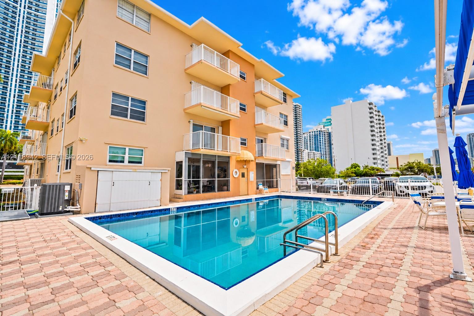 Rectangular swimming pool with tiled coping, surrounded by a brick paver deck, featuring multi-story building exterior with private balconies and glass-enclosed ground level space