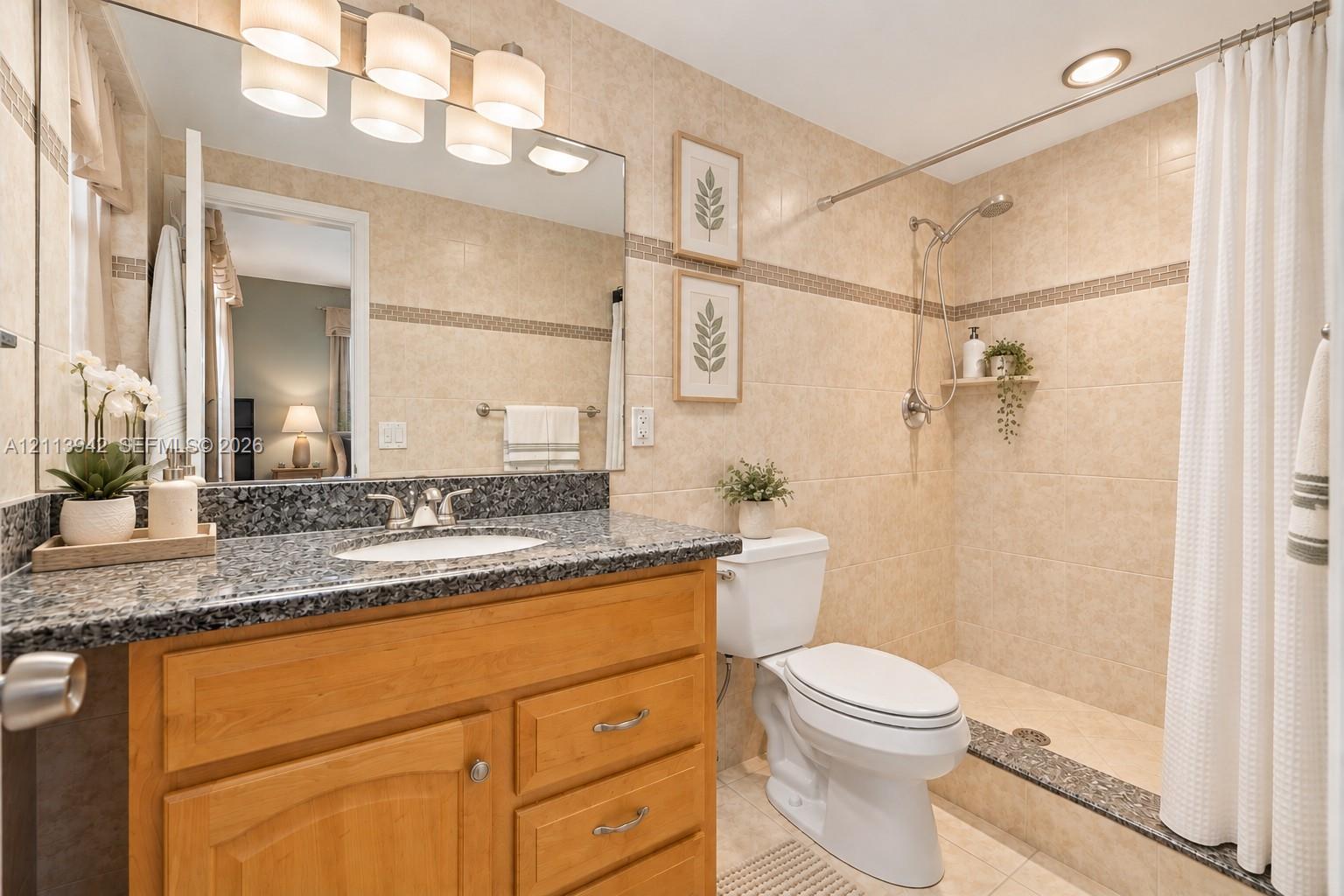Bathroom featuring a wood-finish vanity with a stone-top counter, undermount sink, multi-light fixture, and a tiled shower stall with a built-in shelf