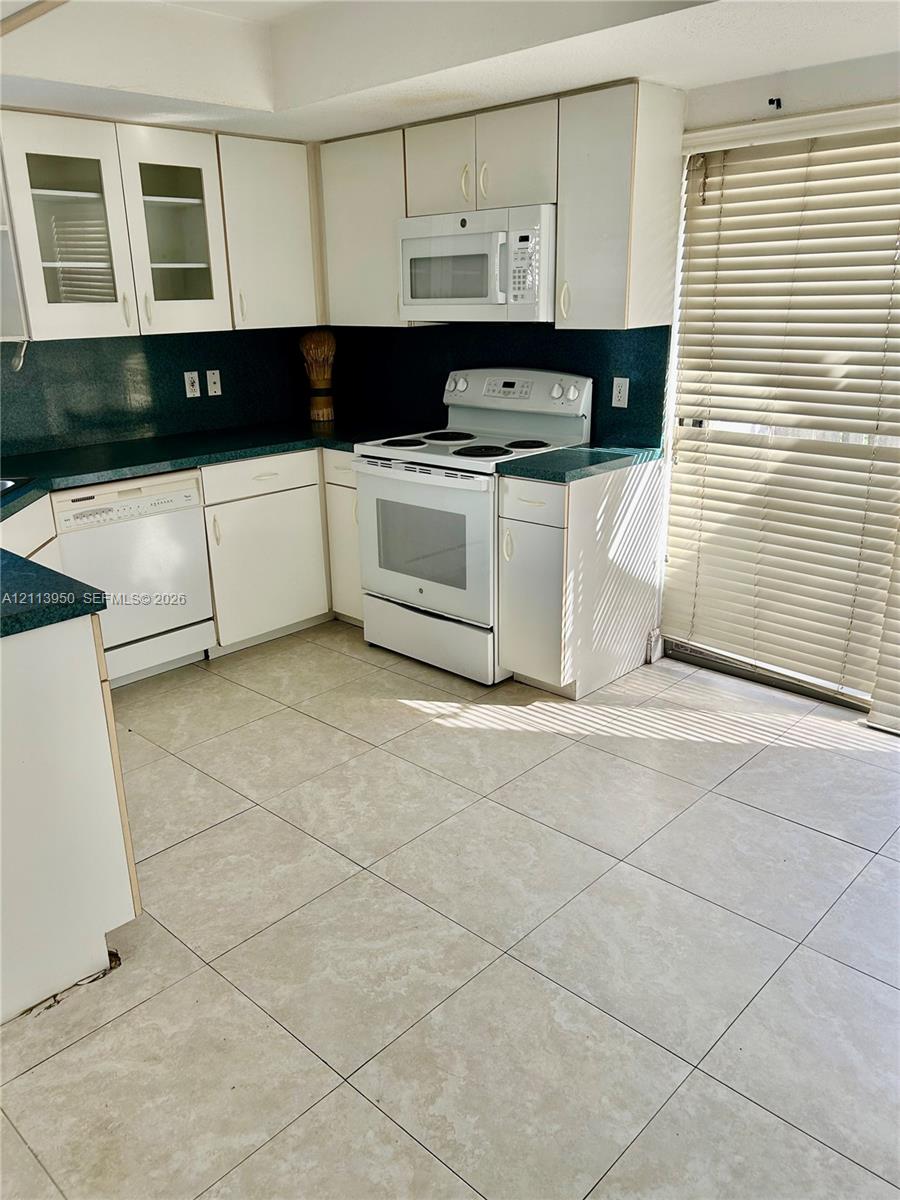 Kitchen featuring white cabinetry with glass-front uppers, solid surface countertops, square tile flooring, and integrated white appliances Kitchen featuring white cabinetry with glass-front uppers, solid surface countertops, square tile flooring, and integrated white appliances