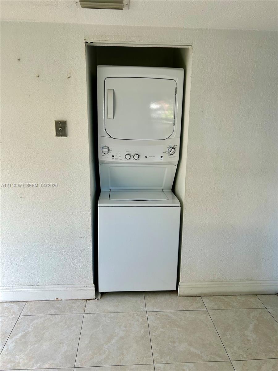 Stacked white laundry unit set within a recessed wall alcove, featuring a top-mounted dryer, bottom-loading washer, and neutral ceramic tile flooring