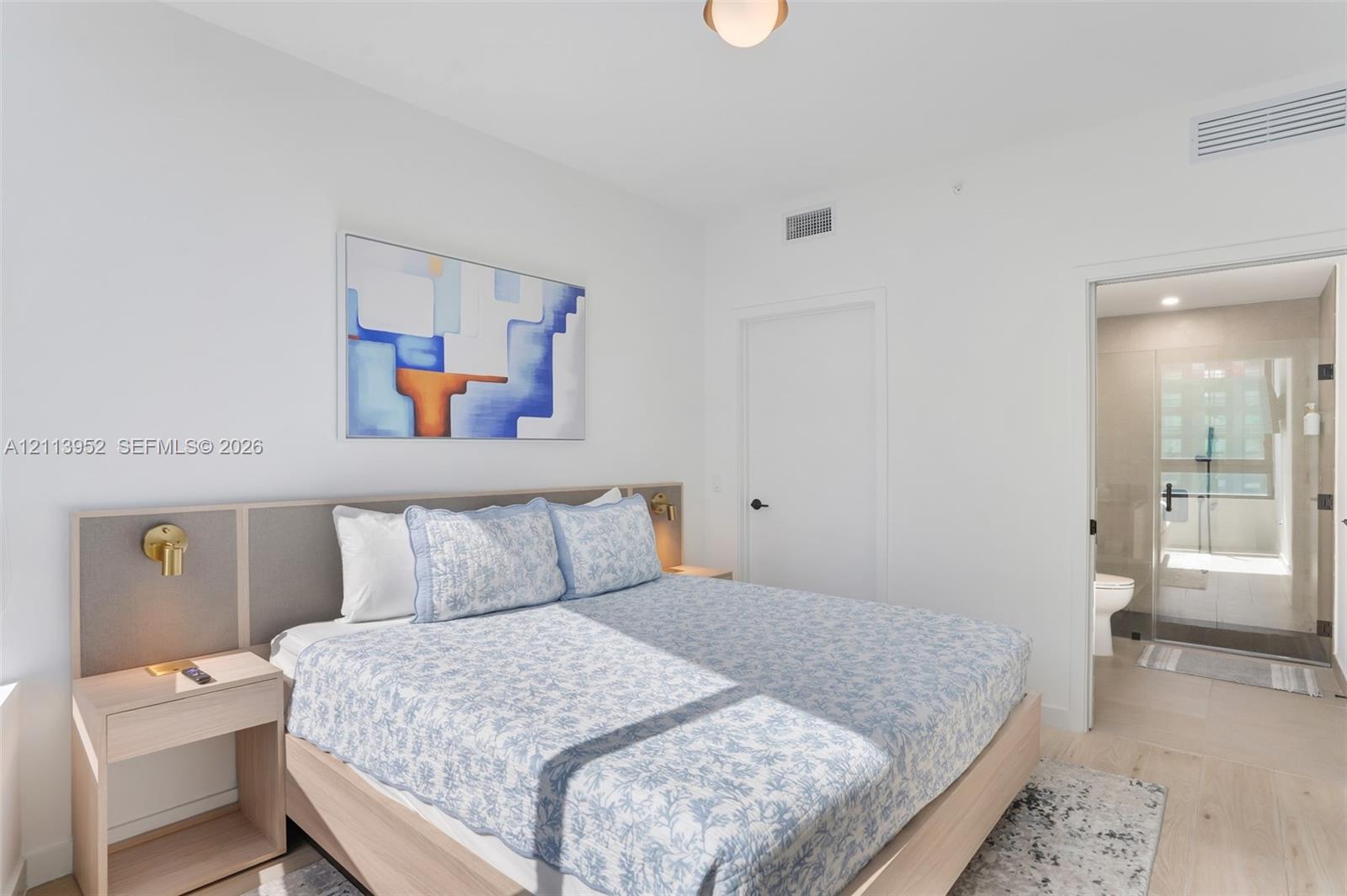 Neutral-toned bedroom featuring a built-in wood-finish headboard with integrated bedside tables, brass wall-mounted light fixtures, light-toned flooring, and a glass-enclosed shower stall