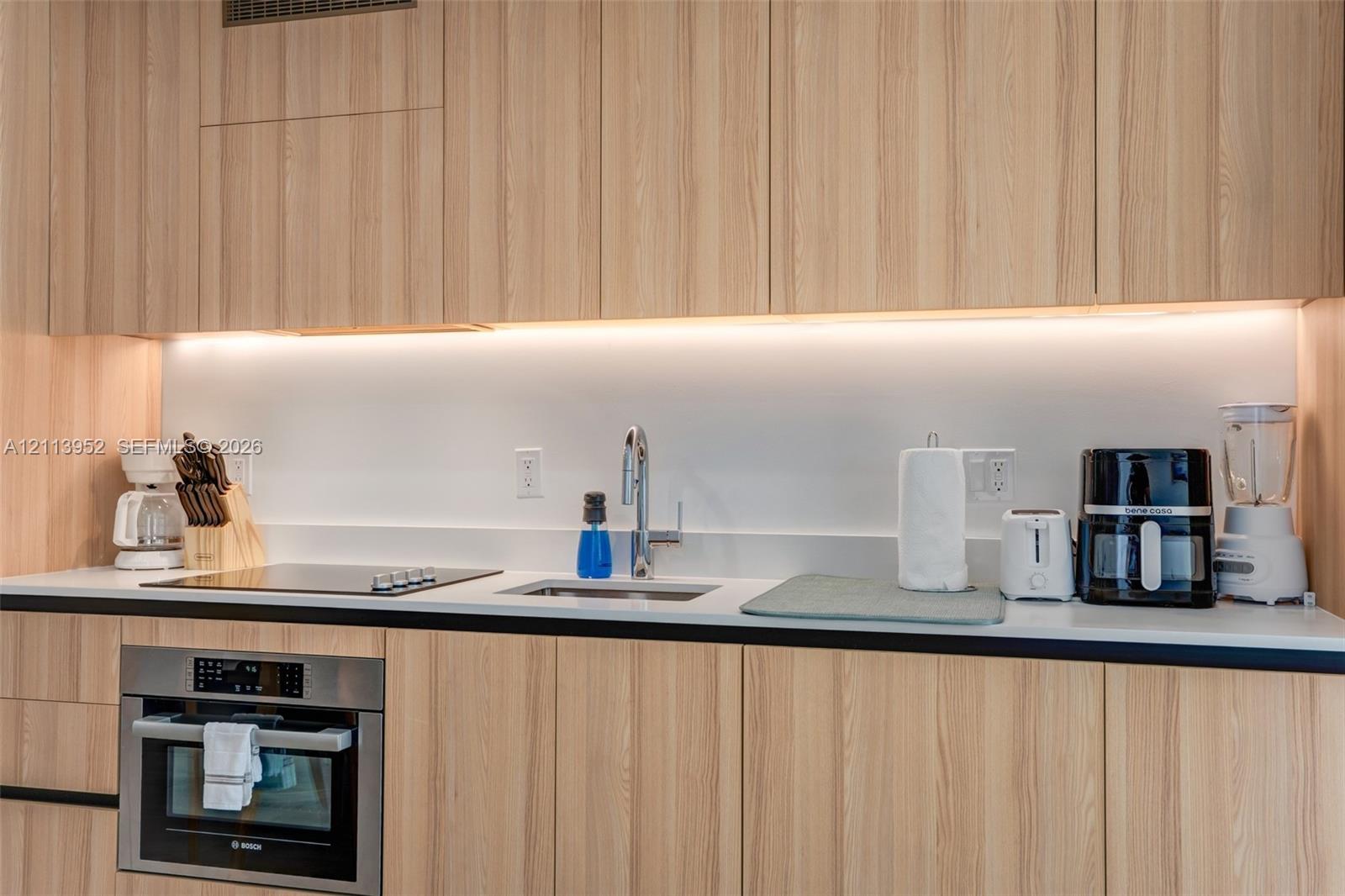 Kitchen featuring light wood-finish cabinetry, solid surface countertops, a flat-surface cooktop, an integrated stainless steel wall oven, and under-cabinet task lighting