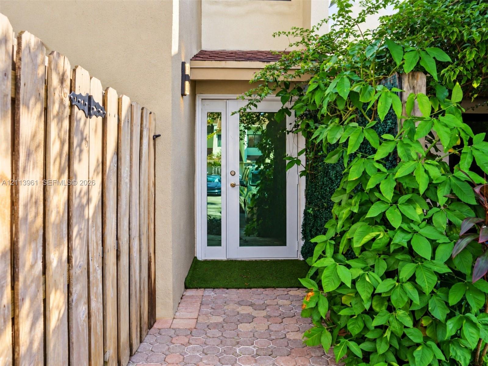 Entrance featuring a white-frame glass door with a sidelight, geometric paver walkway, vertical wood-plank privacy fencing, and exterior wall-mounted lighting Entrance featuring a white-frame glass door with a sidelight, geometric paver walkway, vertical wood-plank privacy fencing, and exterior wall-mounted lighting