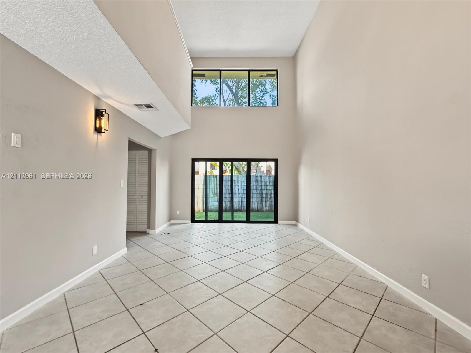 High-ceilinged interior featuring large-format tile flooring, a multi-pane upper window, sliding glass doors, and wall-mounted sconce lighting High-ceilinged interior featuring large-format tile flooring, a multi-pane upper window, sliding glass doors, and wall-mounted sconce lighting