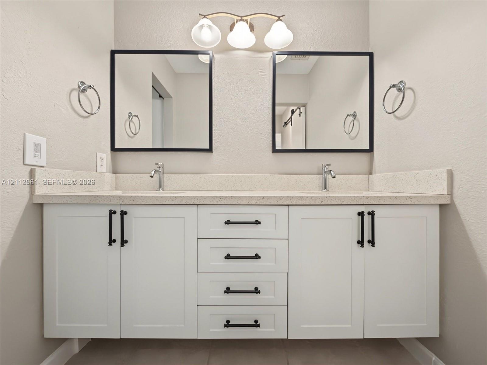 Dual-sink vanity featuring white cabinetry, solid surface countertop, matte black hardware, square mirrors with black frames, and a three-bulb light fixture