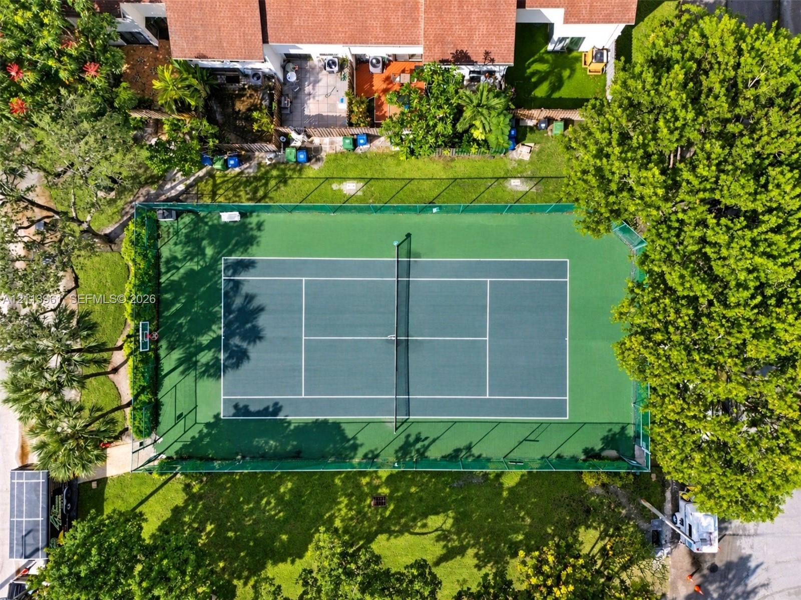 Full-size tennis court featuring a perimeter fence, basketball hoop, and surrounding manicured lawn with mature trees