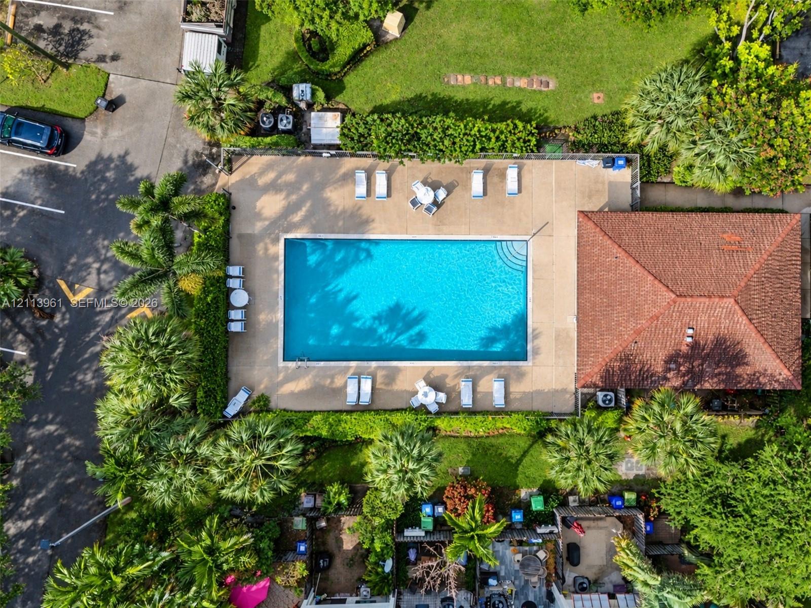 Rectangular swimming pool with surrounding concrete deck, terracotta-tiled roof structure, and perimeter landscaping featuring mature trees