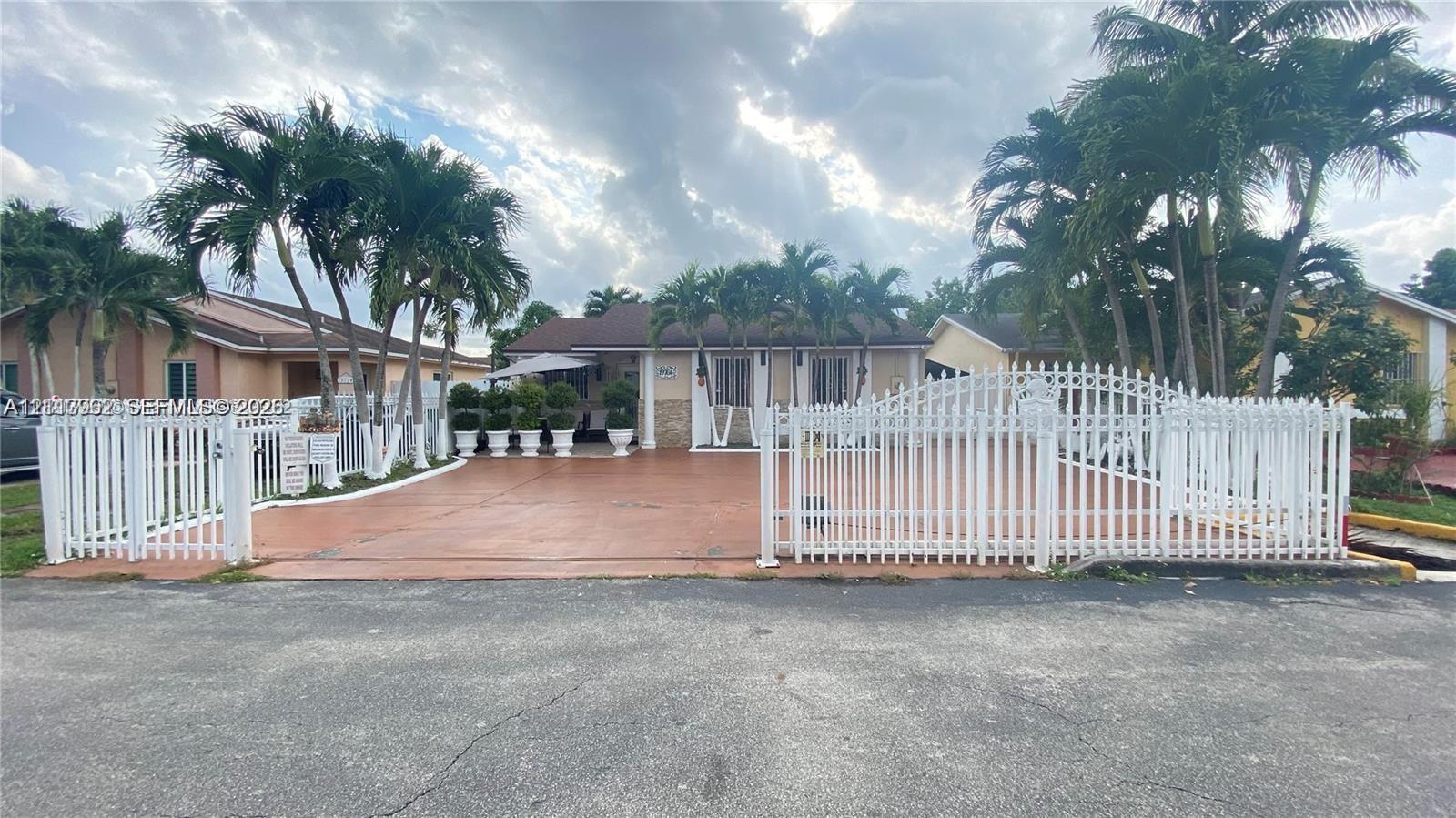 Exterior facade featuring a paved driveway, a white perimeter fence with gated access, and mature palm trees Exterior facade featuring a paved driveway, a white perimeter fence with gated access, and mature palm trees