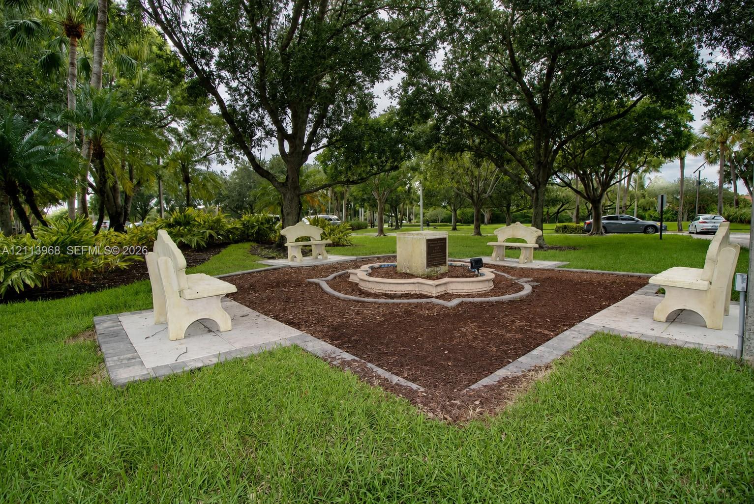 Landscaped grounds featuring stone benches, a central stone pedestal, mulched garden beds, and mature shade trees