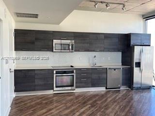 Kitchen featuring dark wood-grain cabinetry, quartz countertops, white hexagonal tile backsplash, stainless steel appliances, and exposed concrete ceilings with track lighting