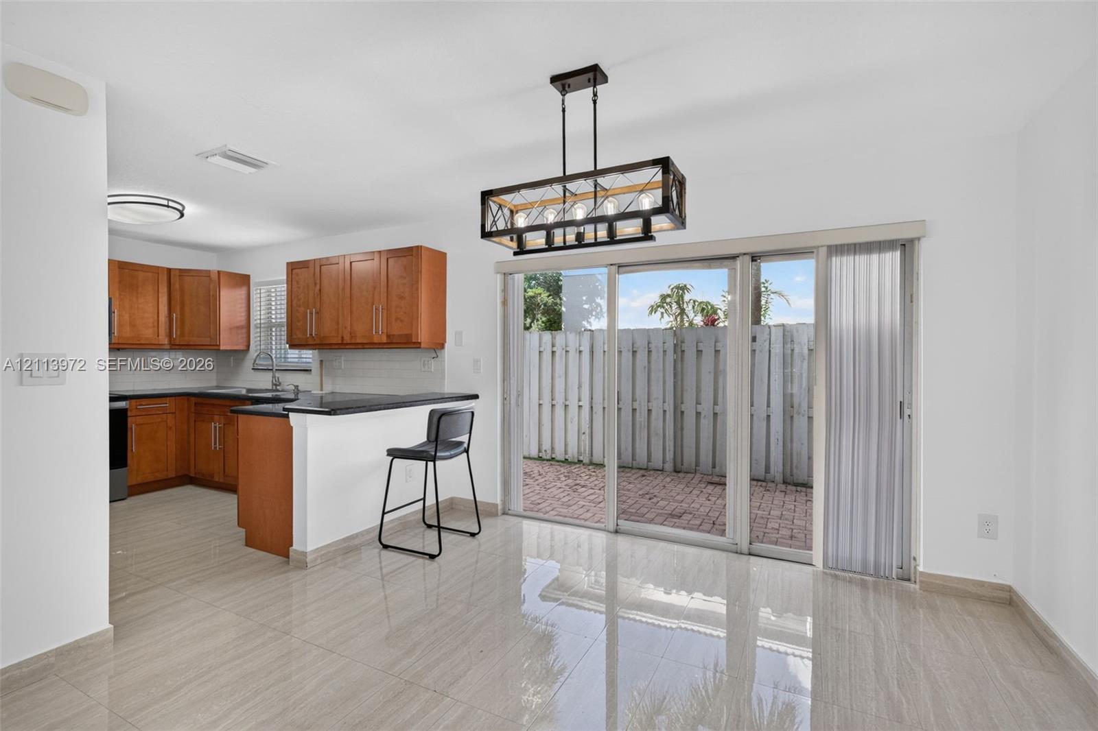 Kitchen featuring wood cabinetry, a breakfast bar with stone-finish countertops, tile flooring, a geometric multi-light chandelier, and sliding glass doors opening to a brick-paved outdoor space Kitchen featuring wood cabinetry, a breakfast bar with stone-finish countertops, tile flooring, a geometric multi-light chandelier, and sliding glass doors opening to a brick-paved outdoor space