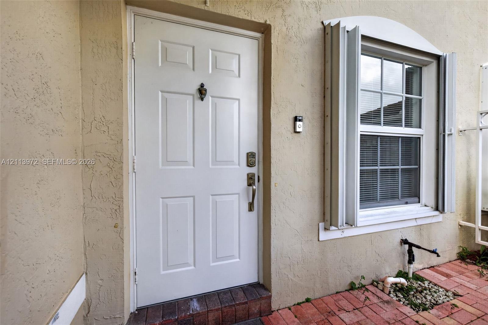 Exterior entryway featuring a six-panel white door, arched window with metal shutters, textured stucco siding, and a red brick patio Exterior entryway featuring a six-panel white door, arched window with metal shutters, textured stucco siding, and a red brick patio