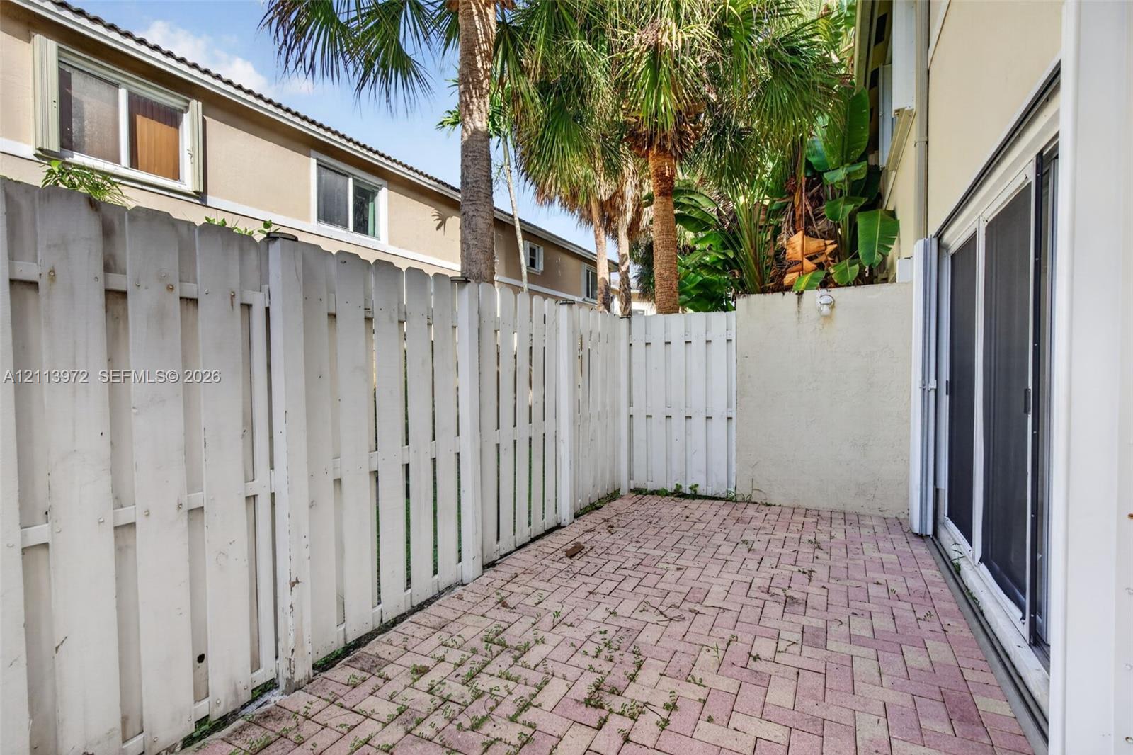 Private rear patio featuring herringbone-patterned brick pavers, enclosed by a white privacy fence and mature palm trees