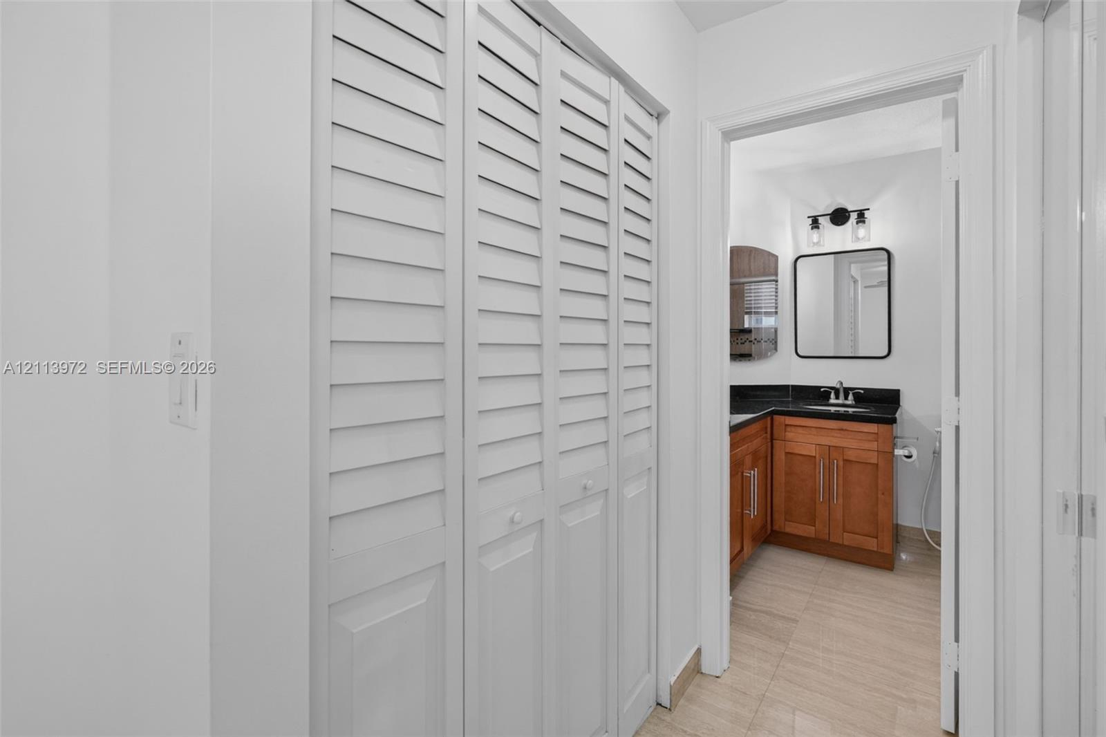 White louvered-panel closet doors featuring wood-finish cabinetry, a dark stone countertop, an integrated rectangular vanity sink, and a black-framed rectangular wall mirror