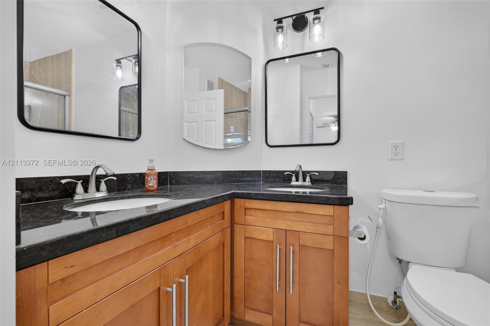 Dual-vanity cabinetry featuring natural wood finish, dark stone countertops, undermount sinks, rectangular framed mirrors, and industrial-style wall-mounted lighting