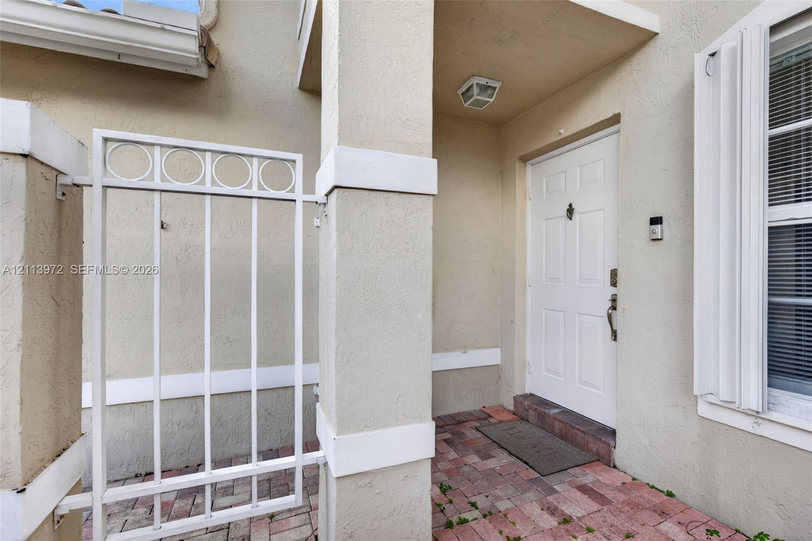 Covered entryway featuring a brick paver floor, white decorative metal gate, paneled front door with a metal knocker, and exterior shutter accents