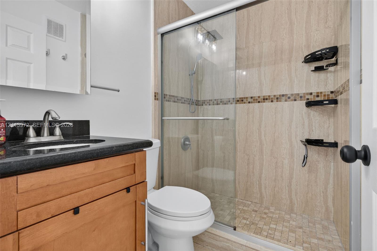 Bathroom featuring a wood-finish vanity with a dark stone countertop, an integrated sink, a glass-enclosed shower with tiled walls, and a mosaic tile accent border