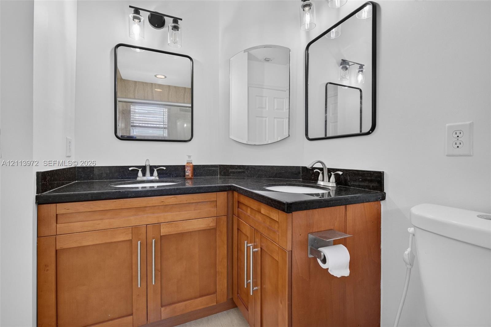 Dual-sink vanity with wood-finish cabinetry, dark stone countertops, dual rectangular wall mirrors with black frames, and three-light vanity fixtures