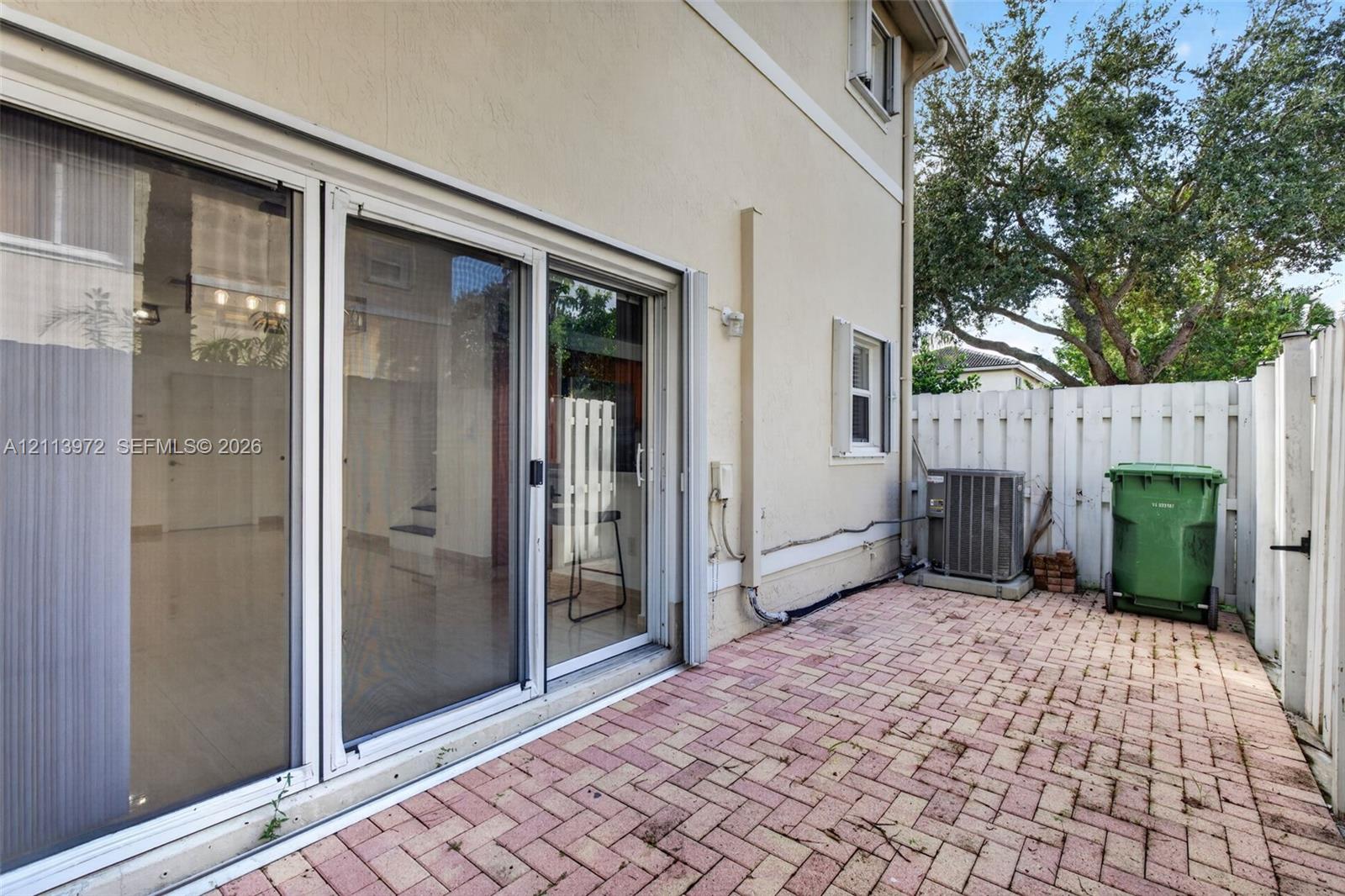 Exterior brick-paver patio featuring a multi-panel sliding glass door system, stucco siding, and a white privacy fence