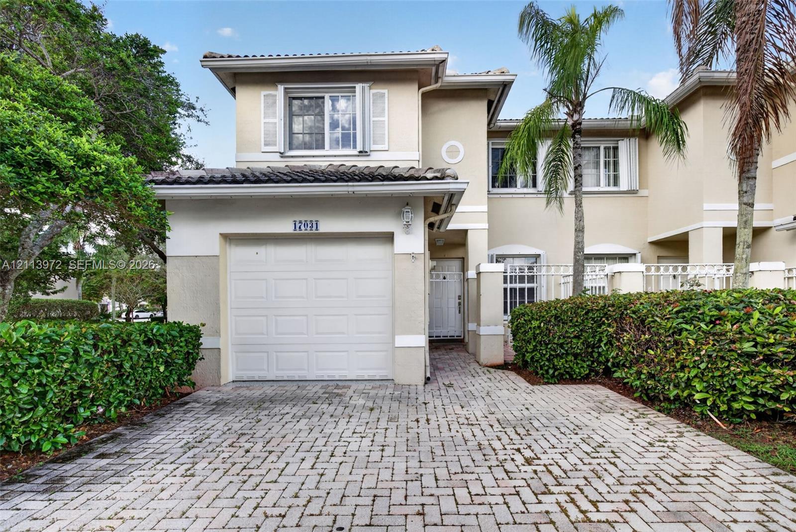 Two-story stucco exterior featuring a tiled roof, attached single-car garage, brick-paver driveway, and manicured perimeter landscaping