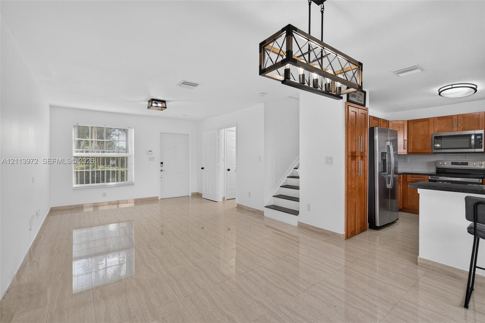 Large-format tile flooring, kitchen featuring wood cabinetry and stainless steel appliances, rectangular pendant lighting with metal frame, and staircase with dark-finish treads Large-format tile flooring, kitchen featuring wood cabinetry and stainless steel appliances, rectangular pendant lighting with metal frame, and staircase with dark-finish treads