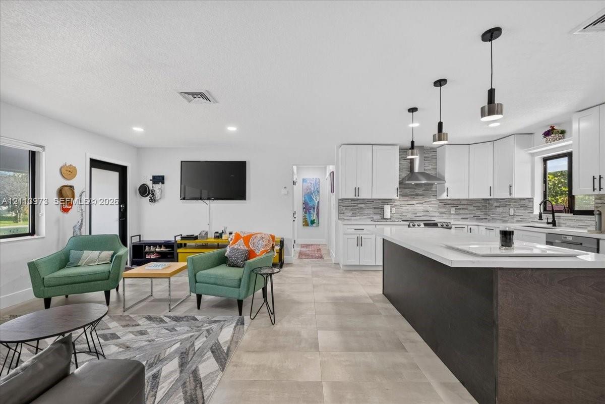 Kitchen featuring white cabinetry, an expansive island with a contrasting dark base, quartz countertops, a stone tile backsplash, and contemporary pendant lighting, set over large-format neutral tile flooring Kitchen featuring white cabinetry, an expansive island with a contrasting dark base, quartz countertops, a stone tile backsplash, and contemporary pendant lighting, set over large-format neutral tile flooring