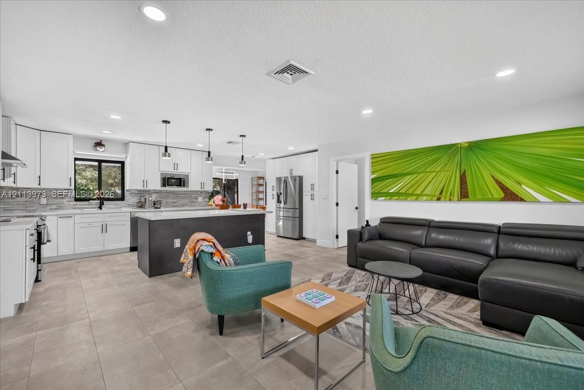 Open-concept kitchen featuring white shaker-style cabinetry, a contrasting dark central island, stone-tiled backsplash, stainless steel appliances, and large-format neutral floor tiling Open-concept kitchen featuring white shaker-style cabinetry, a contrasting dark central island, stone-tiled backsplash, stainless steel appliances, and large-format neutral floor tiling
