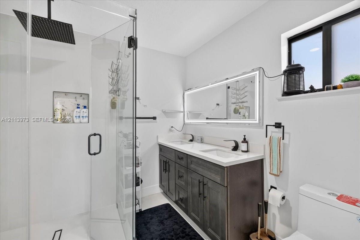 Double vanity featuring a light-toned countertop, dark-finished cabinetry, an illuminated mirror, a glass-enclosed shower with a rainfall showerhead, and a built-in recessed wall niche