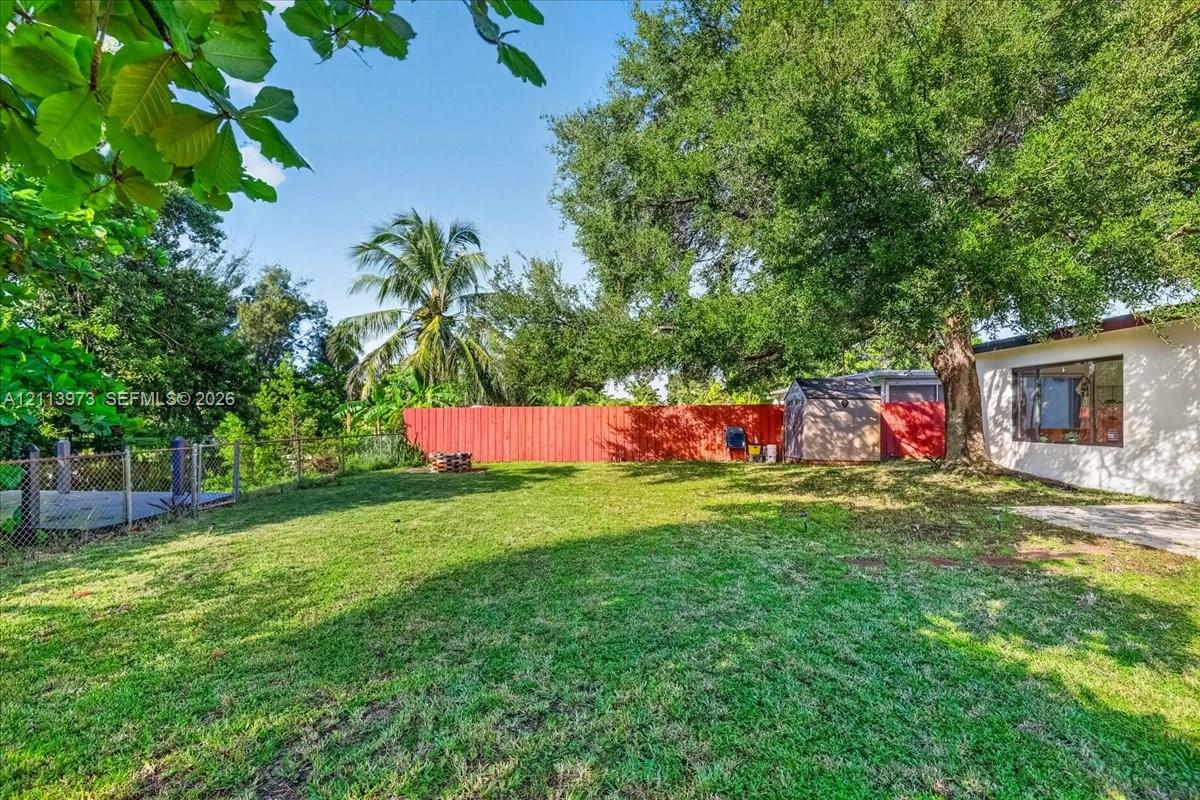 Spacious backyard featuring a manicured lawn, mature trees, red perimeter fencing, and concrete patio