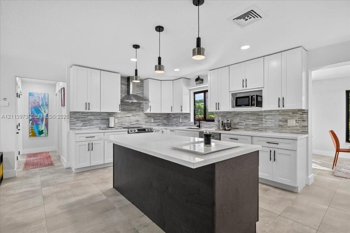 Kitchen featuring white shaker-style cabinetry, a central island with a contrasting dark finish, white solid-surface countertops, multi-toned horizontal tile backsplash, and stainless steel appliances