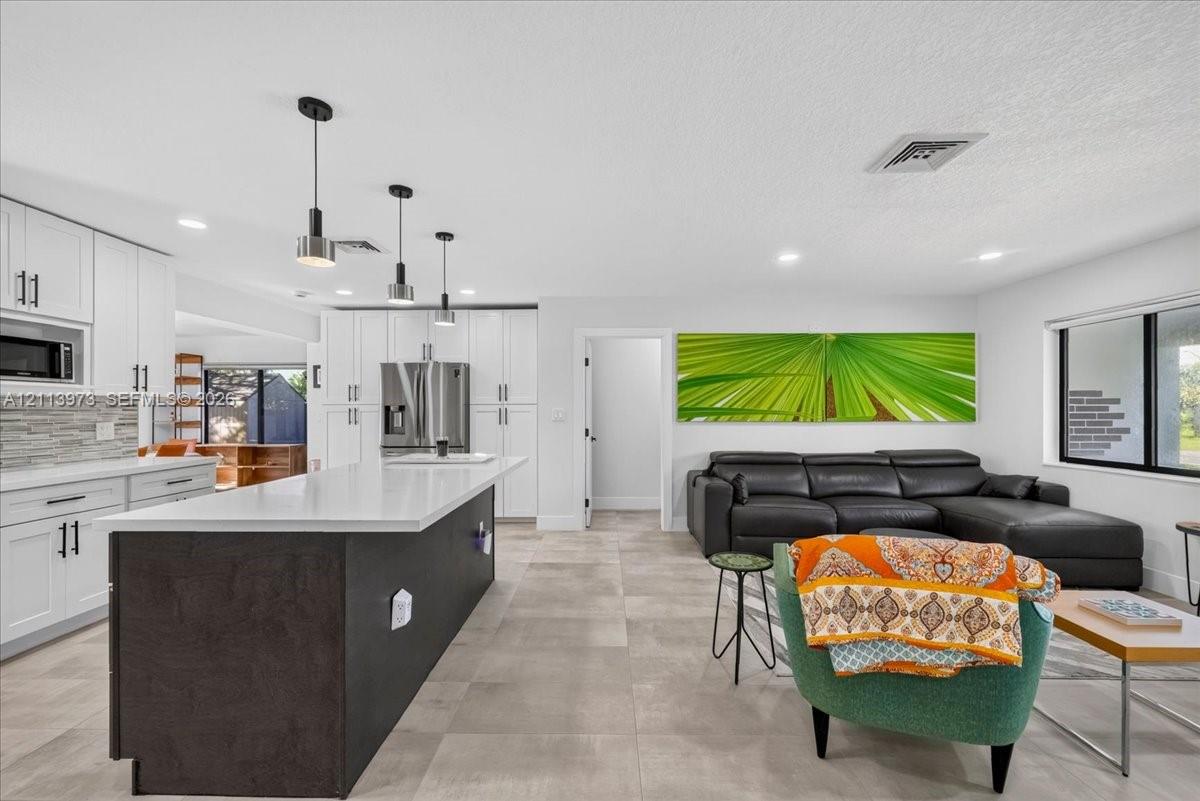 Kitchen featuring white shaker-style cabinetry, an expansive center island with a dark finish, quartz countertops, and a stacked stone backsplash, complemented by large-format tile flooring throughout